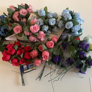 May include: Assortment of artificial rose bouquets in various colors, including pink, red, blue, and purple. Each bouquet features multiple small roses with green leaves and wire stems. Some bouquets have tags attached.