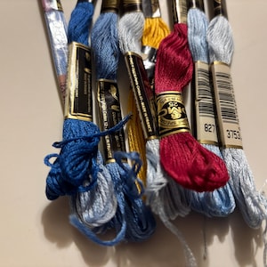 May include: A collection of embroidery floss in various colors, including shades of blue, yellow, and red. The floss is bundled and labeled with product information. The image shows a close-up of the spools, highlighting the different colors and textures of the thread.