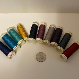May include: A collection of colorful spools of thread, including shades of blue, teal, gold, and red. The spools are arranged in a semi-circle, with a quarter coin in the center. The thread is labeled "Gütermann".