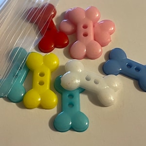 May include: A collection of colorful plastic bone-shaped buttons. The buttons are in shades of blue, yellow, pink, red, and white. Each button has two holes for sewing.