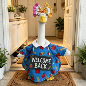 May include: A white goose figurine wearing a blue dress with a pattern of red apples, pencils, and the letters ABC. The goose wears a large yellow and pink bow, round glasses, and holds a black sign with the white text WELCOME BACK.