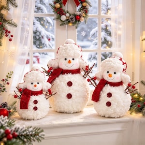 May include: Three plush white snowman figures with fuzzy textures, wearing red and white patterned trapper hats and red scarves. Each snowman features a carrot nose, black button eyes, and twig arms adorned with small red berries.