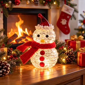 Könnte beinhalten: Eine Schneemann-Figur aus Glas mit rissiger Textur, die von innen beleuchtet wird. Sie trägt einen roten Strickschal, eine rote Mütze mit weißem Rand sowie drei rote runde Knöpfe, schwarze Augen und eine gelbe Nase als weihnachtliche Dekoration.