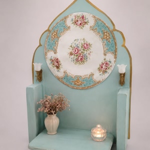 May include: A light blue wooden wall shelf with a pointed arch top and gold trim. A large circular plate with a floral pattern is mounted on the back panel. Two small wall sconces flank the plate. A small vase with dried flowers and a glass candle holder sit on the shelf.