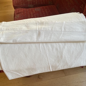 May include: A folded white bed sheet featuring a textured fabric surface and a horizontal row of decorative hemstitch detailing along the top edge. The textile is opaque and displays a consistent off-white tone across its entire visible surface area.