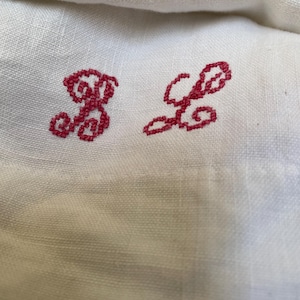 Puede incluir: Una tela blanca que presenta dos letras bordadas con hilo rojo oscuro. Las letras están cosidas en un estilo de escritura cursiva con bucles. La tela tiene una textura tejida visible y el bordado está centrado en el material.