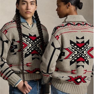 May include: A beige knit sweater featuring a shawl collar and a geometric pattern in red, black, and white. The design includes arrow motifs and bold angular shapes across the chest and back, finished with ribbed cuffs and a ribbed hemline.