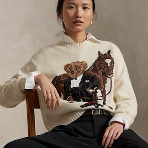 Op de afbeelding: Een crèmekleurige gebreide trui met lange mouwen en een centrale intarsia-afbeelding van een teddybeer in een zwart smokingkostuum naast een bruin paard. De trui heeft een geribbelde ronde hals, boorden en zoom en wordt gedragen over een wit overhemd.