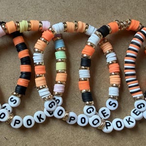 May include: Five circular bracelets made of colorful polymer clay heishi beads and gold-toned spacers. Each bracelet features white round beads with black lettering spelling out the phrase GO POKES, arranged in various color patterns including orange, black, white, and pastel tones.