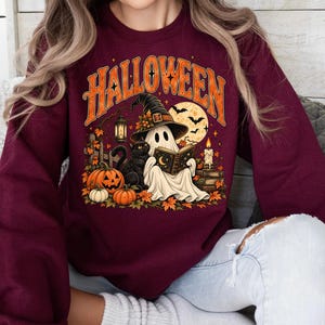 May include: A maroon long-sleeve sweatshirt featuring a graphic print with the text HALLOWEEN in orange. The design includes a ghost wearing a witch hat reading a book, a black cat, pumpkins, a lantern, a candle, and a full moon.