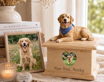 Puede incluir: Una urna de madera clara para mascotas con una figura de golden retriever con un pañuelo azul en la parte superior. El frente de la urna tiene una abertura en forma de corazón para una foto y el texto grabado "Run free, Rocky" debajo.
