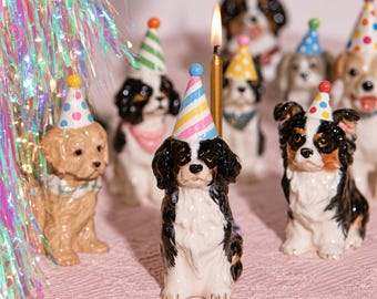 May include: A collection of ceramic dog figurines, each wearing a colorful party hat. The dogs feature various breeds and coat colors, including black, white, and tan. A single gold birthday candle stands lit among the figurines.
