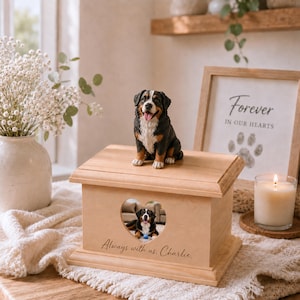 May include: A light-colored wooden pet urn featuring a sculpted Bernese Mountain Dog figurine on top. The front has a heart-shaped cutout displaying a photo of a dog and the engraved text "Always with us. Charlie." The urn has a rectangular base.