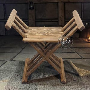 May include: A folding wooden stool with a natural brown wood grain finish. The stool features an X-shaped base and two upward-angled side supports that form a seat. A circular logo with the text Alexander Hamilton is visible on the right side.