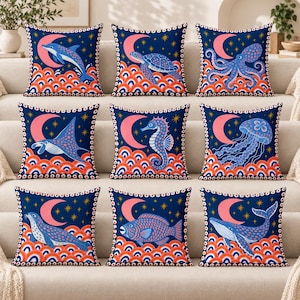 Puede incluir: Un conjunto de nueve cojines cuadrados con un fondo azul oscuro y un patrón repetitivo de olas naranjas, una luna creciente rosa y estrellas amarillas. Cada cojín muestra un animal marino diferente: delfín, tortuga, pulpo, raya, caballito de mar, medusa, foca, pez y ballena.