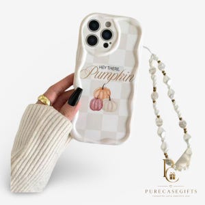 May include: A white wavy-edged phone case featuring a light gray checkered pattern, the text "HEY THERE. Pumpkin" in brown script, and three watercolor-style pumpkins in orange, pink, and white. A beaded phone strap with white and gold beads is attached.
