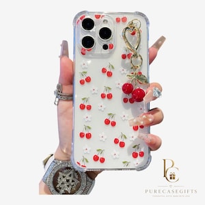 Peut inclure: Une coque de téléphone transparente ornée d'un motif répétitif de petites cerises rouges et de fleurs blanches. Un charm en métal doré en forme de cœur avec deux perles cerises rouges pendantes est fixé en haut à droite.