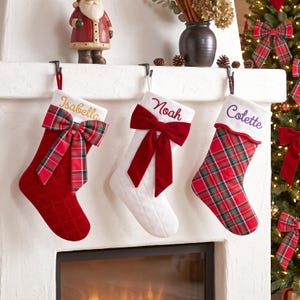 Peut inclure: Trois chaussettes de Noël suspendues à une cheminée. La première est rouge avec un nœud écossais et le nom Isabella. La deuxième est blanche avec un nœud en velours rouge et le nom Noah. La troisième est écossaise avec un revers blanc et le nom Colette.