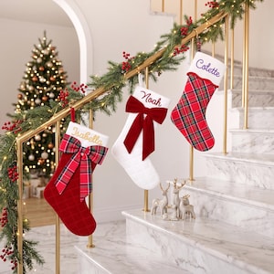 Peut inclure: Trois chaussettes de Noël suspendues à une rampe d'escalier dorée. L'une est rouge avec un nœud écossais et le nom Isabella, une blanche avec un nœud en velours rouge et le nom Noah, et une rouge écossaise avec un revers blanc et le nom Colette.