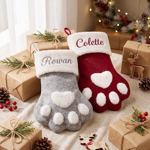 May include: Two paw-shaped Christmas stockings with white sherpa cuffs. The grey stocking features the name Rowan embroidered in grey, and the red stocking features the name Colette embroidered in red. Both have white sherpa heart and toe pads.