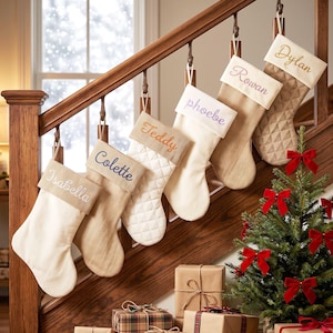 Peut inclure: Cinq chaussettes de Noël suspendues à une rampe d'escalier en bois. Les chaussettes présentent des revers brodés avec les prénoms Isabella, Colette, Teddy, Phoebe, Rowan et Dylan. Elles sont en tissu crème et en toile de jute beige matelassée.