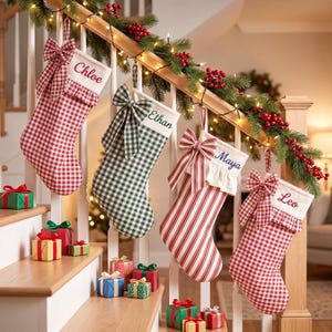 Peut inclure: Quatre chaussettes de Noël suspendues à une rampe d'escalier en bois. Trois chaussettes présentent des motifs vichy rouges ou verts, tandis qu'une autre a des rayures verticales rouges. Chaque chaussette a un revers blanc avec un nom.