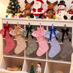 Peut inclure: Six chaussettes de Noël suspendues avec des motifs vichy variés en rouge, beige, vert, rose, bleu et noir. Chaque chaussette présente un revers à volants, un nœud en velours assorti et un prénom brodé sur le revers.
