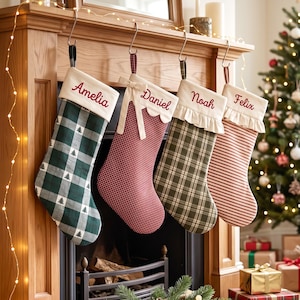 Op de afbeelding: Vier gepersonaliseerde kerstsokken hangend aan een houten schoorsteenmantel. De eerste is groen geruit met boompjes, de tweede rood geblokt met een strik, de derde olijfgroen geruit met ruches en de vierde rood gestreept met ruches.