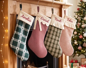 May include: Four personalized Christmas stockings hanging from a wooden mantel. The first is green plaid with trees, the second is red gingham with a bow, the third is olive plaid with ruffles, and the fourth is red striped with ruffles.