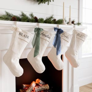 Peut inclure: Quatre chaussettes de Noël en tissu blanc cassé suspendues en rangée. Chaque chaussette présente un revers brodé d'un prénom : BLAIR, Amelia, Brooks ou JULIETTE. Les chaussettes sont ornées de rubans noués blanc, vert sauge et bleu marine.