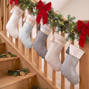 Op de afbeelding: Vijf gepersonaliseerde kerstsokken in wit, lichtblauw en diverse grijstinten, elk voorzien van een crèmekleurige boord geborduurd met namen zoals Teddy, Isabella, Dylan, Rowan en Noah, geaccentueerd met kwastjes en rode strikken.
