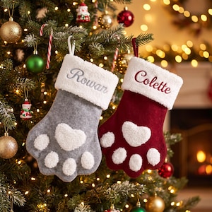 May include: Two paw-shaped Christmas stockings with white sherpa cuffs. The left stocking is gray with the name Rowan embroidered in silver. The right stocking is deep red with the name Colette embroidered in red. Both feature white sherpa pads.