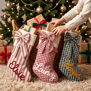 May include: Three fabric Christmas stockings with bows and personalised names. The left stocking has red stripes and the name Elsie. The middle stocking has red gingham and the name Lainey. The right stocking has green gingham and the name Harper.