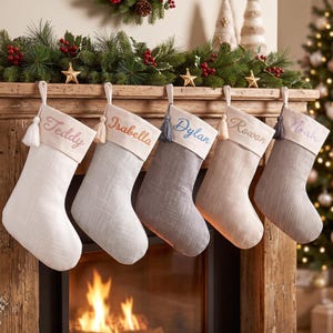 Peut inclure: Cinq chaussettes de Noël en tissu suspendues en rangée, chacune dotée d'un revers couleur crème brodé avec les prénoms Teddy, Isabella, Dylan, Rowan et Noah. Les chaussettes ont des corps texturés dans des tons blanc, gris clair, gris foncé et beige.