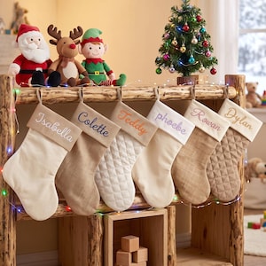 Peut inclure: Six chaussettes de Noël personnalisées suspendues à un support en bois, chacune portant un nom brodé sur le revers : Isabella, Colette, Teddy, Phoebe, Rowan et Dylan. Les chaussettes varient en texture, avec des tissus lisses et matelassés.