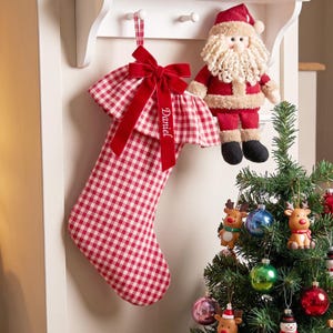 Peut inclure: Une chaussette de Noël à carreaux vichy rouges et blancs avec un haut à volants et un nœud en velours rouge foncé portant le prénom Daniel brodé en fil blanc. La chaussette est suspendue à un crochet blanc près d'une poupée Père Noël.