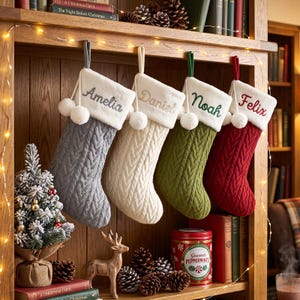 May include: Four hanging Christmas stockings featuring a cable knit texture and a white plush cuff. Each stocking is personalised with embroidered names: Amelia in grey, Daniel in beige, Noah in green, and Felix in red. Each cuff has two white pom-poms.