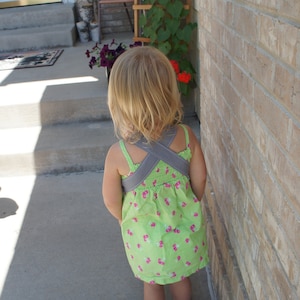 May include: A young girl wearing a green and pink floral dress with a gray criss-cross strap back. She is standing on a concrete patio with a brick wall behind her.