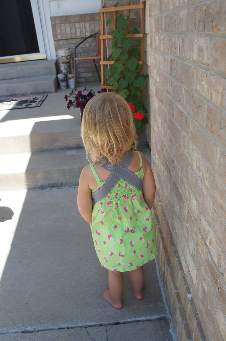 May include: A young girl wearing a green and pink floral dress with a gray criss-cross strap back. She is standing on a concrete patio with a brick wall behind her.