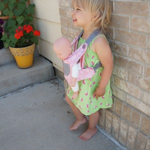 May include: A young girl wearing a green and pink floral dress is carrying a baby doll in a pink and gray baby carrier. The girl is standing next to a brick wall.
