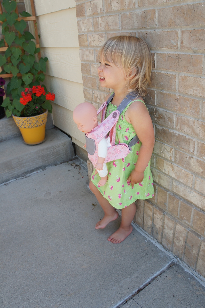 May include: A young girl wearing a green and pink floral dress is carrying a baby doll in a pink and gray baby carrier. The girl is standing next to a brick wall.
