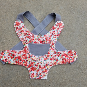 May include: A gray and white baby carrier with a floral pattern. The carrier has a gray pocket on the front and adjustable straps.