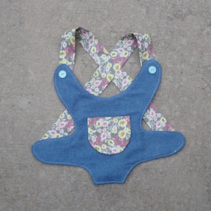 May include: A denim overall for a pet with a floral print fabric strap. The overall has a pocket on the front and two blue buttons on the straps.