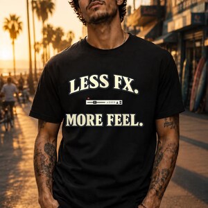Op de afbeelding: Een zwart T-shirt met korte mouwen voorzien van de witte tekst LESS FX. boven een afbeelding van een horizontale audioschuifregelaar en de tekst MORE FEEL. in witte letters. Het shirt heeft een ronde hals en een standaard pasvorm van katoen.