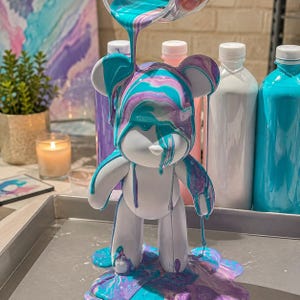Può includere: Una statuetta di orso bianco ricoperta da una colata di vernice acrilica turchese e viola. La vernice gocciola sulle orecchie, sulla testa e sul corpo dell'orso, accumulandosi alla base. Sullo sfondo sono visibili diverse bottiglie di vernice.