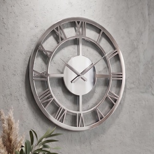 Könnte beinhalten: Eine kreisförmige Wanduhr mit metallisch silbernem Finish, die ein offenes Rahmendesign mit großen römischen Ziffern aufweist. Die Uhr hat eine solide mittlere Scheibe mit zwei dünnen, geraden Zeigern aus Metall.