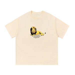 Op de afbeelding: Een crèmekleurig T-shirt met korte mouwen en een afbeelding van een liggende leeuw met donkere manen. Onder de leeuw staat de tekst: The wicked flee when no one pursues but the righteous are bold as a lion.