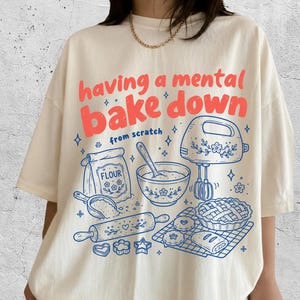 May include: An ivory t-shirt featuring the red text 'having a mental bake down' and blue line art illustrations of a flour bag, mixing bowl, hand mixer, rolling pin, and baked goods with the text 'from scratch' printed on the front.