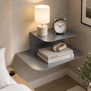 Può includere: Una mensola da parete in metallo a due livelli con finitura grigio opaco. Il ripiano superiore ospita una lampada cilindrica con base in pietra e una sveglia analogica argentata. Il ripiano inferiore ospita due libri impilati AALTO e THE KINFOLK HOME.