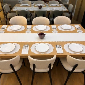 Könnte beinhalten: Ein Esstisch aus hellem Holz für sechs Personen mit weißen, wellenförmigen Platzsets, weißen Tellern, silbernem Besteck und weißen Servietten. Ein langer weißer Tischläufer liegt in der Mitte mit einer Glasschale voller roter Deko.